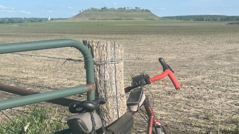Gravel bicycle leaning against a green gate and wooden fence post in front of a plowed field with a hill and silo in the distance, near Greeley, Kansas, on a sunny day.