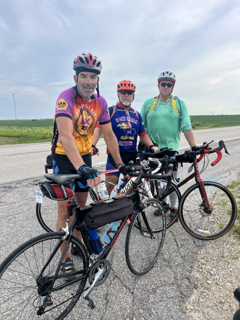 Three cyclists posing in front of a windmill on a sunny day — a trio of road warriors with wind and wheels on their side.