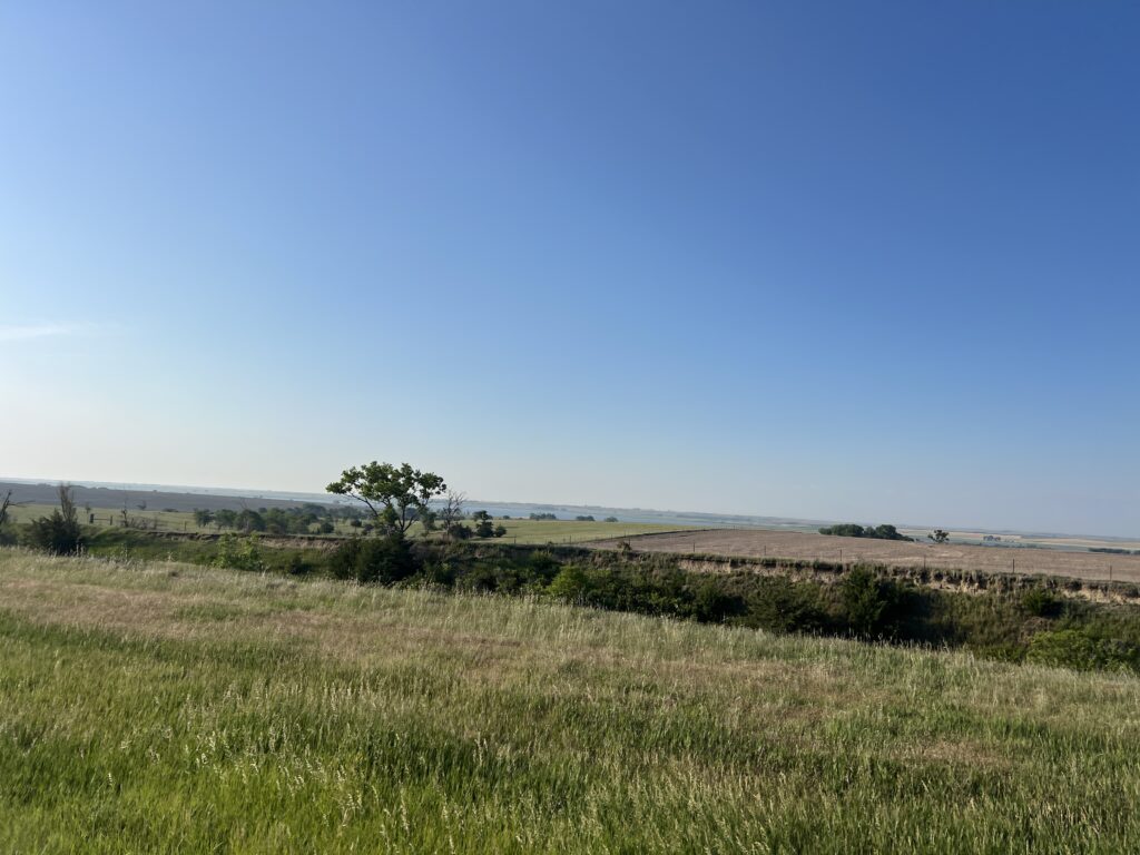 Expansive Kansas countryside with green fields, scattered trees, and a bright blue sky.