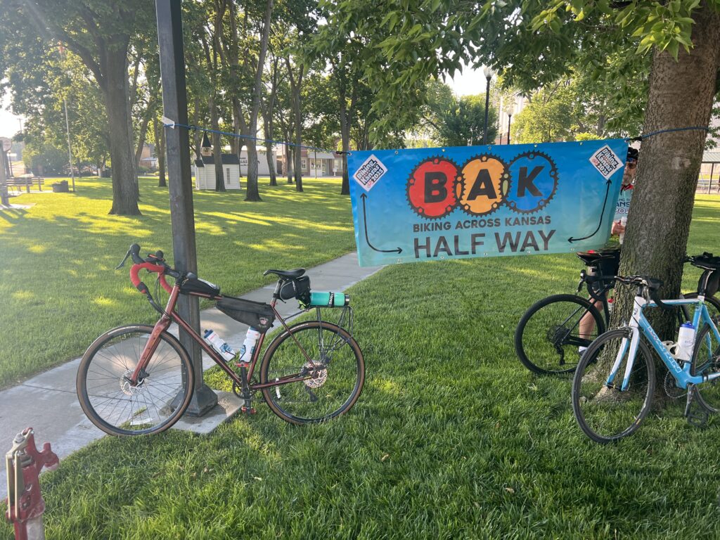 Bicycle parked beside a colorful banner marking the halfway point across Kansas during the Bike Across Kansas 2025 event, with open road and blue skies in the background.