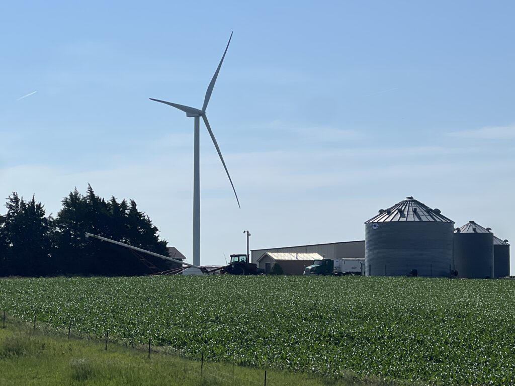 Large windmill standing tall next to a classic farm home, surrounded by open fields under a clear sky.