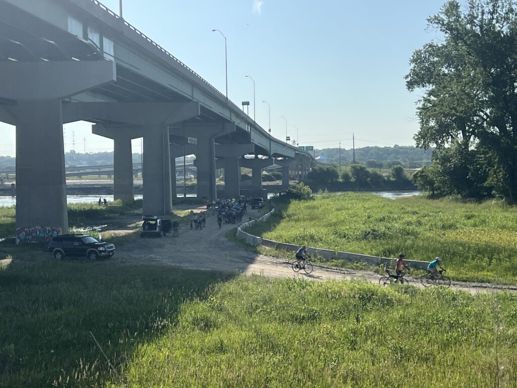 Cyclists dipping their rear tires in the Missouri River beneath a large bridge in Elwood, Kansas, marking the end of the Bike Across Kansas 2025 ride.