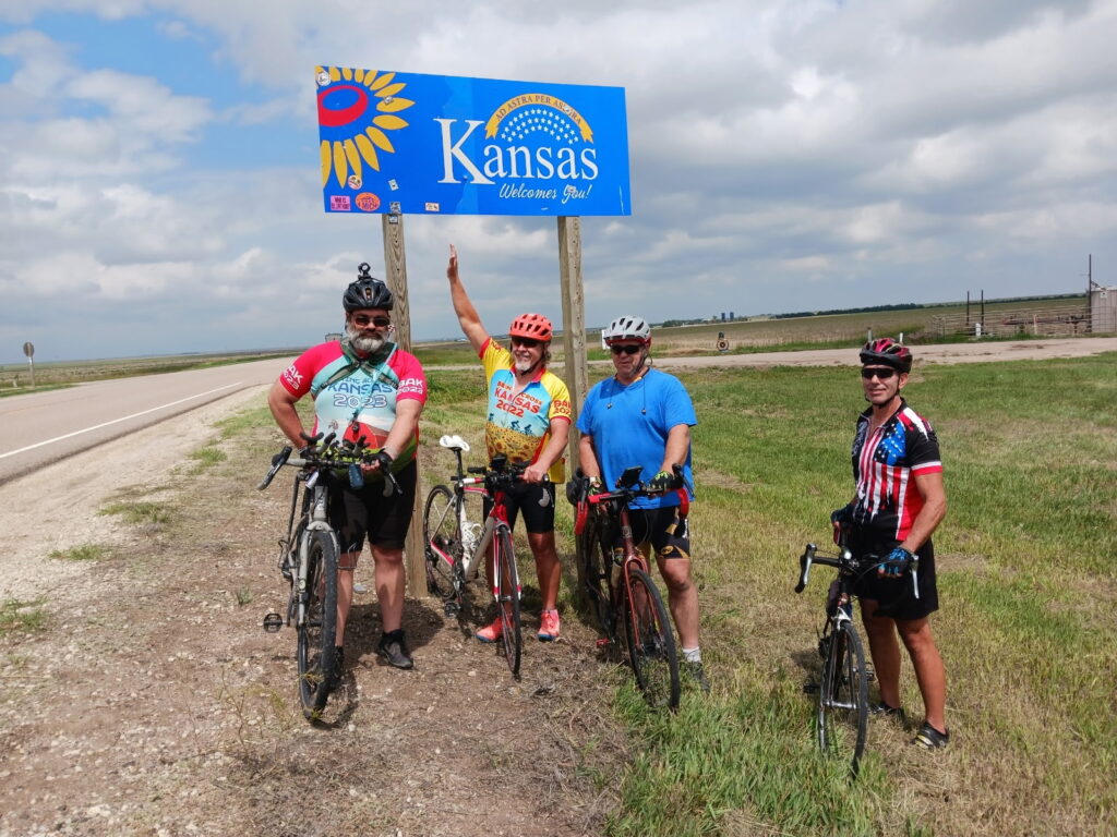 Four cyclists posing beneath the Kansas welcome sign at the Colorado-Kansas state line at the start of Bike Across Kansas.