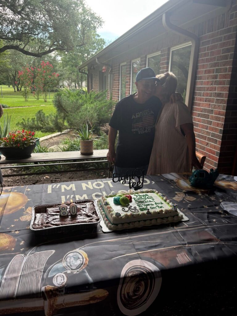 Mom and stepfather standing together in front of the birthday cake, arms around each other, smiling at the camera.