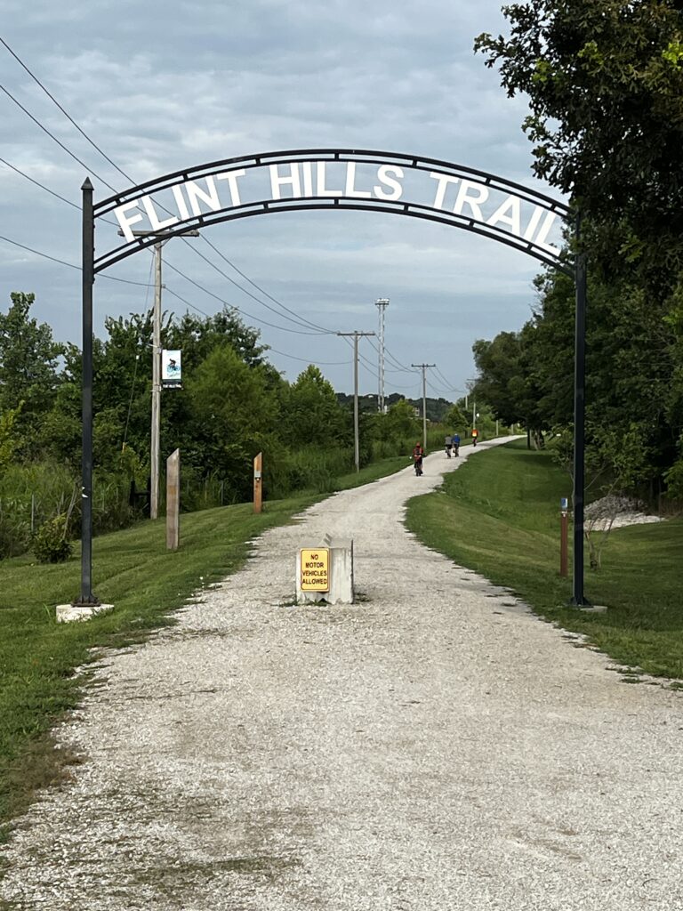 Flint Hills Trail and Sheldon Trail overhead signage at Mile Zero in Osawatomie, Kansas with distant cyclists on the path.