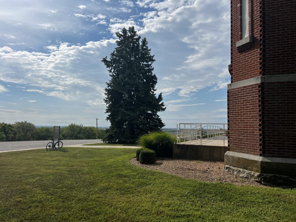 Corner of Sacred Heart Catholic Church with a large cedar tree, looking south across the Kansas countryside under a cloudy sky