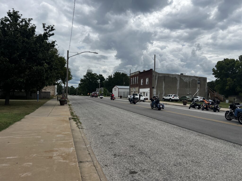 Cyclists and motorcyclists leaving Guy & Mae’s Tavern in Williamsburg, Kansas, bikes and noisy motorcycles on the street, friends sharing smiles and laughter as they head out.
