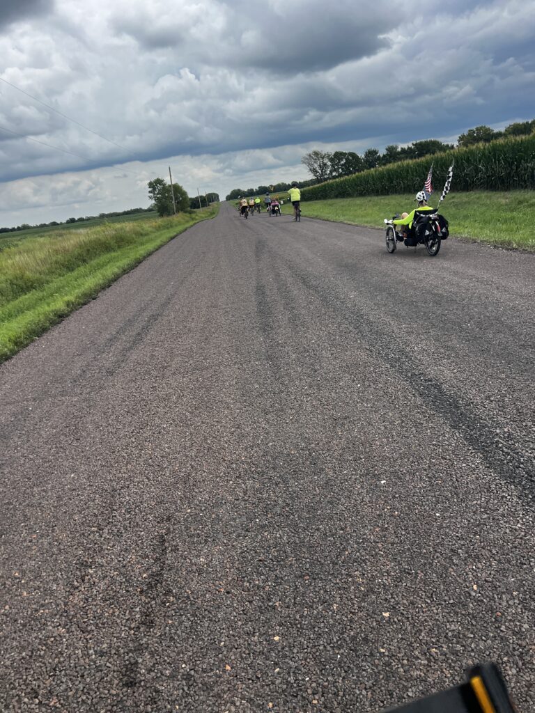 Group of cyclists riding on a chip-and-seal road toward Princeton Trailhead, Kansas. Riders pause as one cyclist deals with rear tire trouble, while friends offer support before continuing