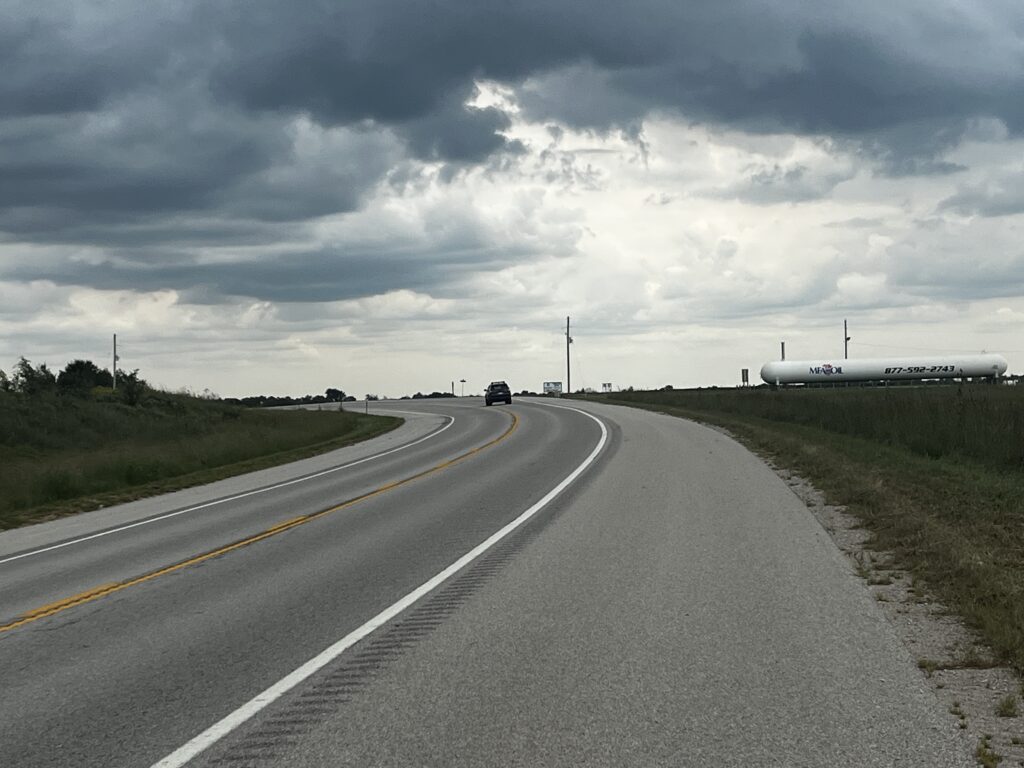 Cyclist riding along a curve on Highway 59 in Kansas, with a wide 12-foot shoulder and dramatic clouds overhead.