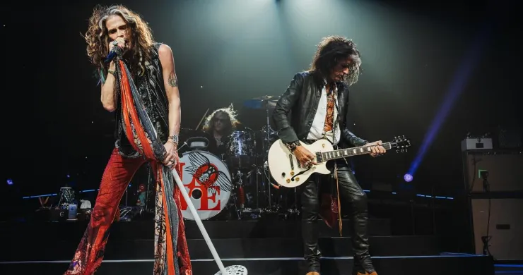 Steven Tyler performing on stage with Aerosmith, striking his signature mic-stand pose, band in full background.