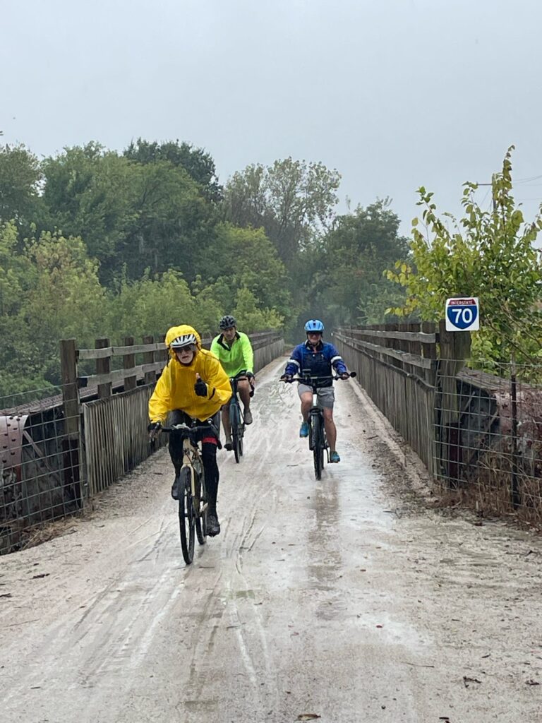 Cyclists riding the Katy Trail just over the I-70 bridge with rain falling and wet ground visible.
