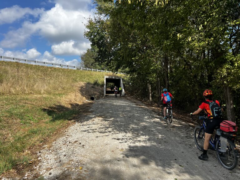 Cyclists on the Katy Trail under Highway 52, trees overhead on a sunny day, ShauneNation adventure.
