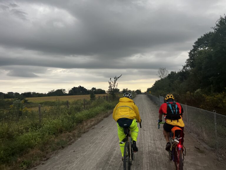 Cyclists riding the Katy Trail in raincoats under dark clouds and steady rain near Clifton, Missouri.