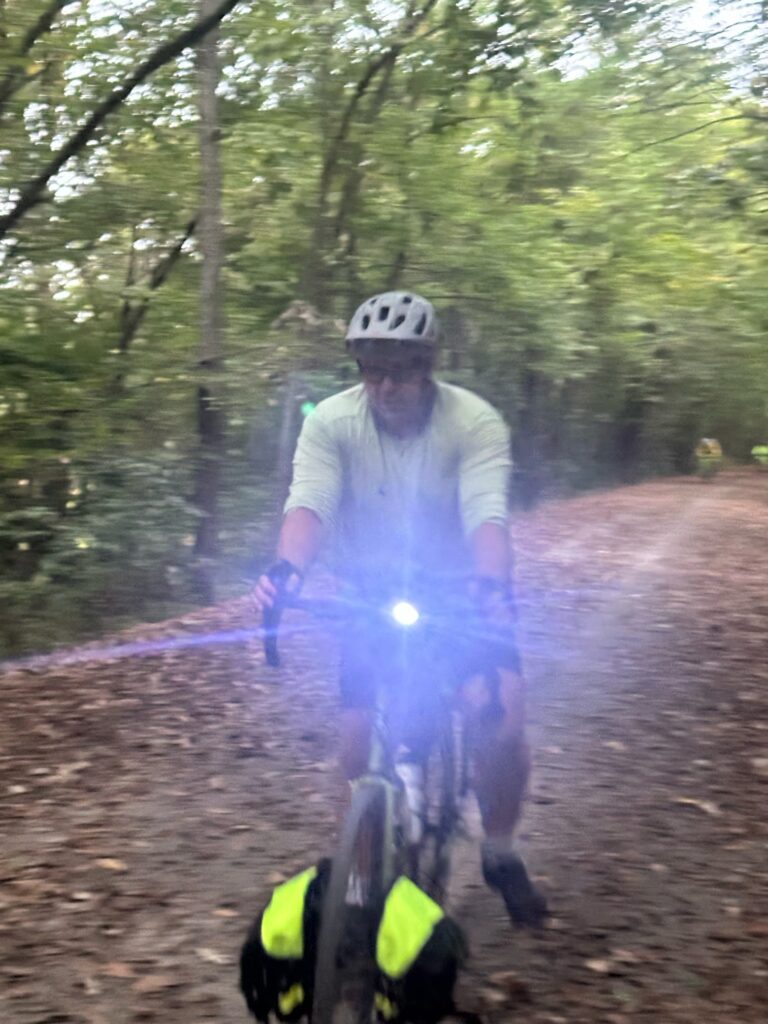 A cyclist leads the group on the Katy Trail near Clifton, Missouri, headlight bright under dark, stormy clouds.