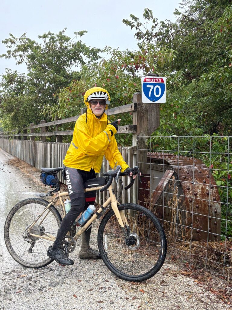 Cyclist named Randy pointing at an Interstate 70 sign while standing on the Katy Trail bridge, celebrating turning 70 this December.