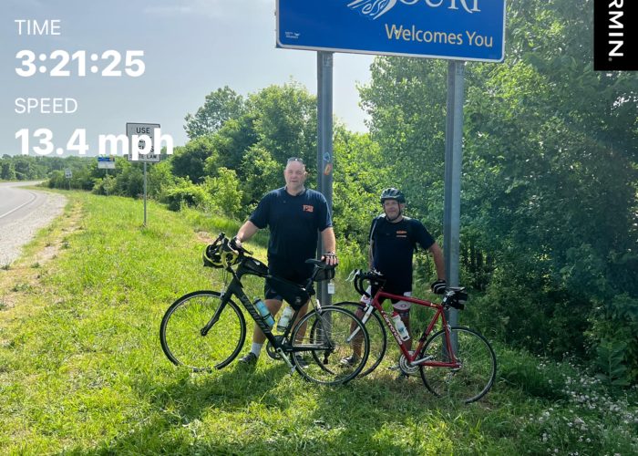 Two cyclists with their bikes standing under the Missouri state line sign, marking the end of Bike Across Kansas.