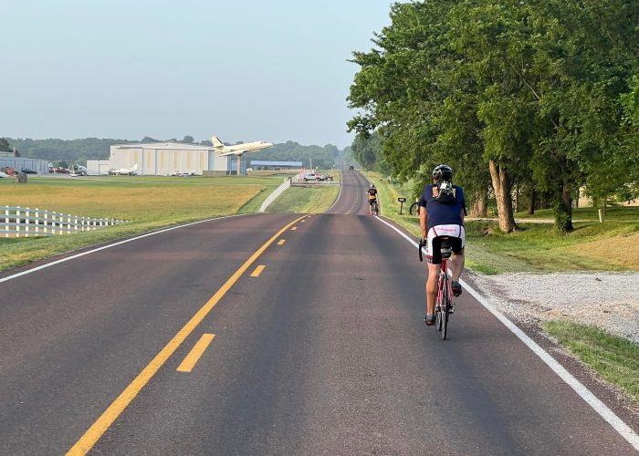 Bicyclists riding on the road in Rantoul, Kansas, passing a Lockheed JetStar mounted above Dodson International Parts aircraft salvage yard during a Bike Across Kansas ride.
